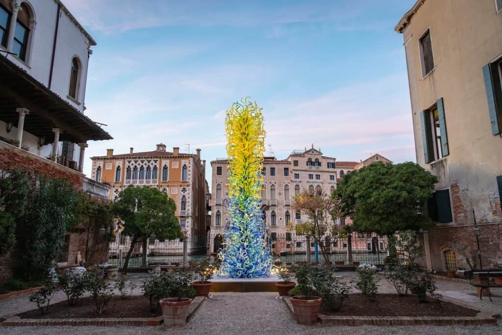 Tall glass sculpture with yellow and blue swirling shapes stands in a garden courtyard, surrounded by historic buildings under a clear sky.