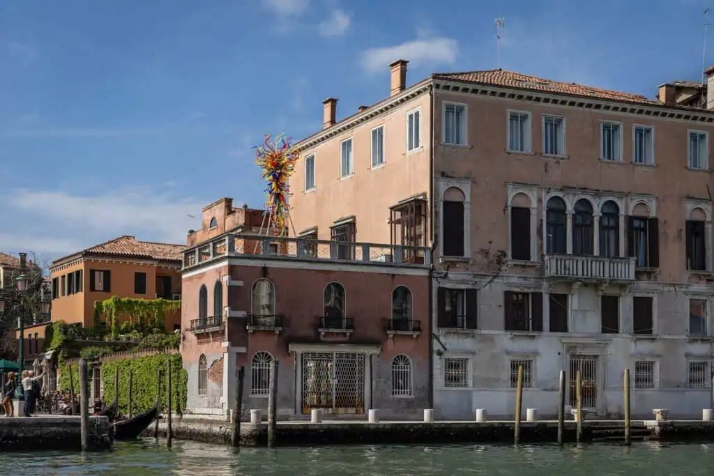 A large ornate sculpture sits on the rooftop of a pink historic building beside a canal in Venice, Italy, with clear skies overhead.