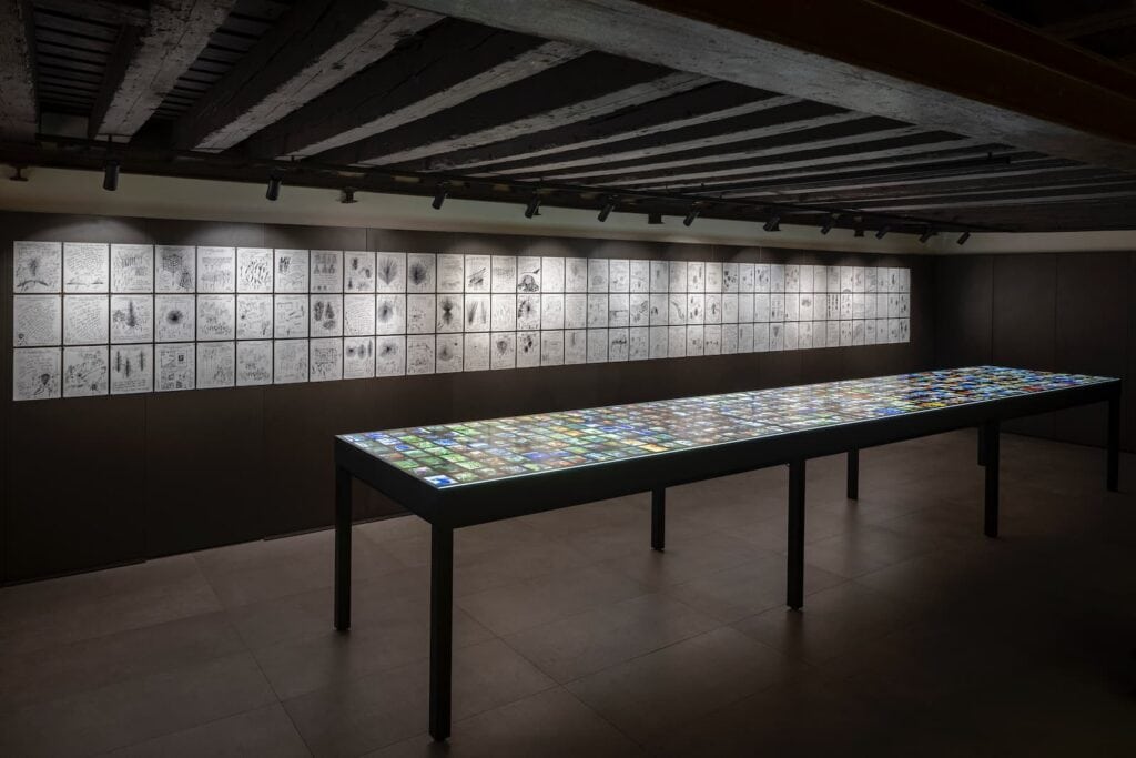 A long, illuminated table displays colorful images, while black-and-white sketches are arranged in a grid on the wall behind, in a dimly lit exhibition space with a wooden ceiling.