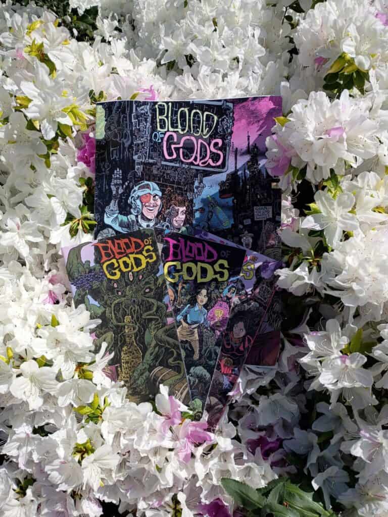 Several "Blood of Gods" comic books are displayed among white and pink flowering bushes outdoors.