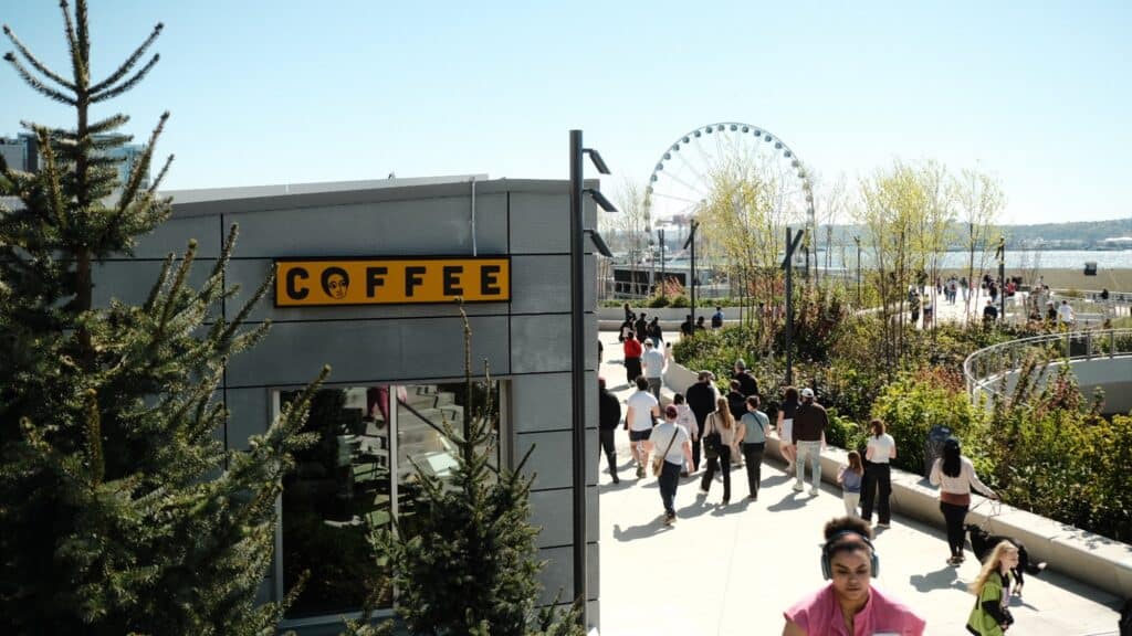 People walk along a path near a modern building with a yellow "COFFEE" sign; a Ferris wheel and waterfront are visible in the background on a sunny day.