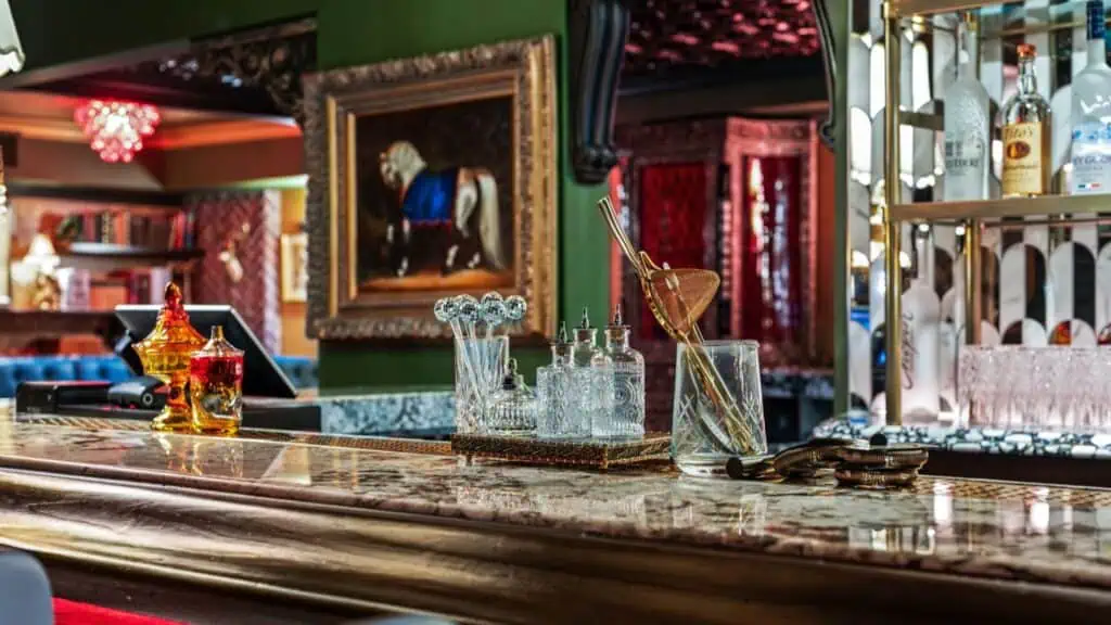 A bar counter displays glassware, cocktail tools, amber jars, and liquor bottles on shelves in the background.