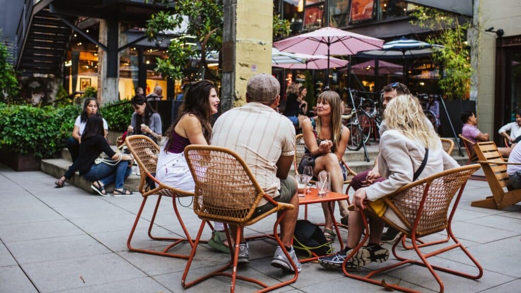 A group of people sit and talk around a small table at an outdoor patio, with shops, plants, and other people visible in the background.