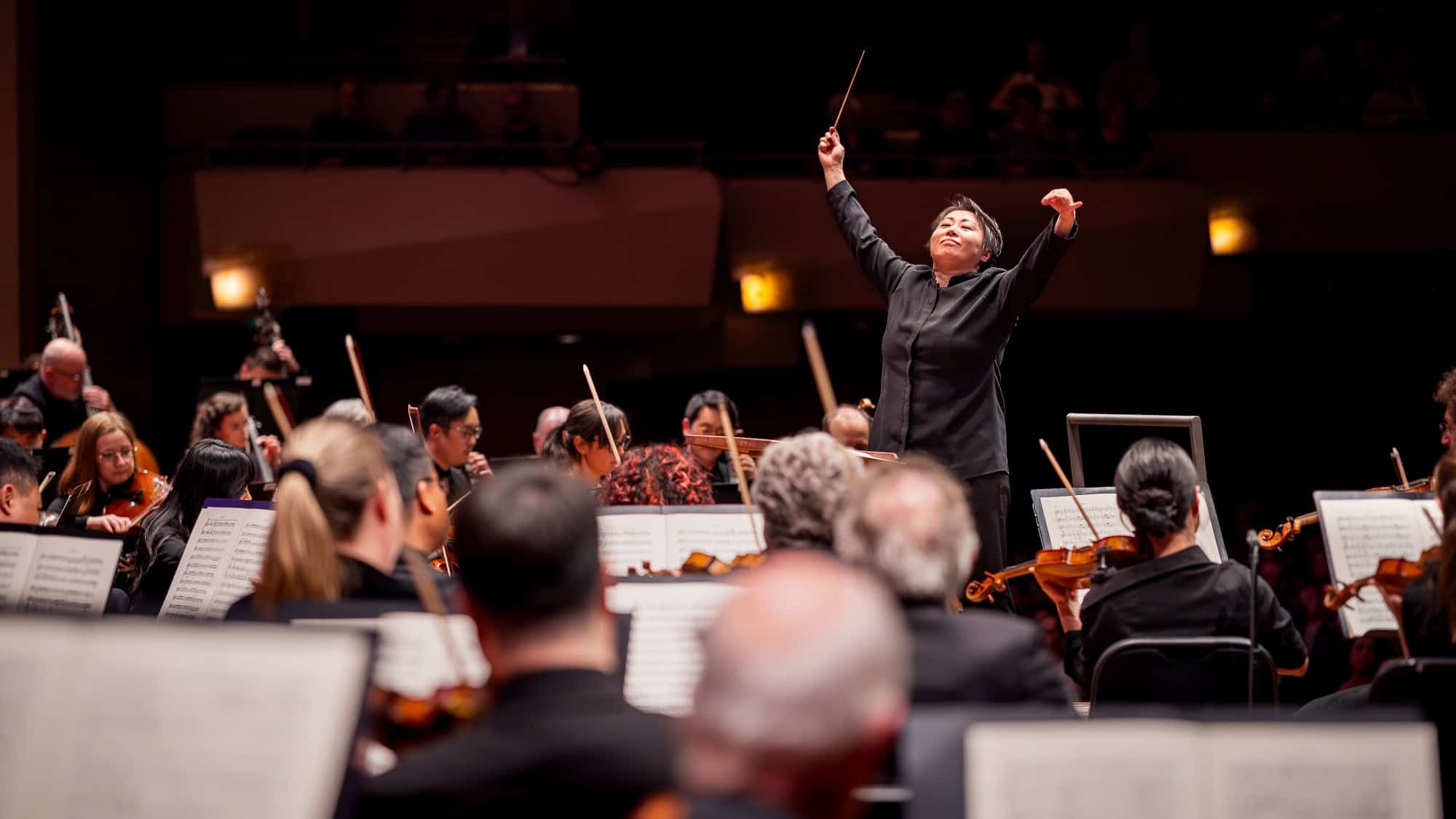 A conductor leads an orchestra during a performance, with musicians playing various string instruments and sheet music visible in the foreground.