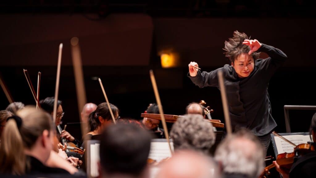 A conductor energetically leads an orchestra during a live performance, with musicians playing string instruments in the foreground.