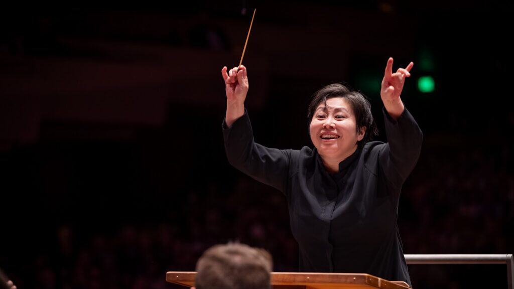 A conductor stands at a podium, holding a baton in one hand and gesturing energetically with both arms, leading an orchestra in a dimly lit concert hall.