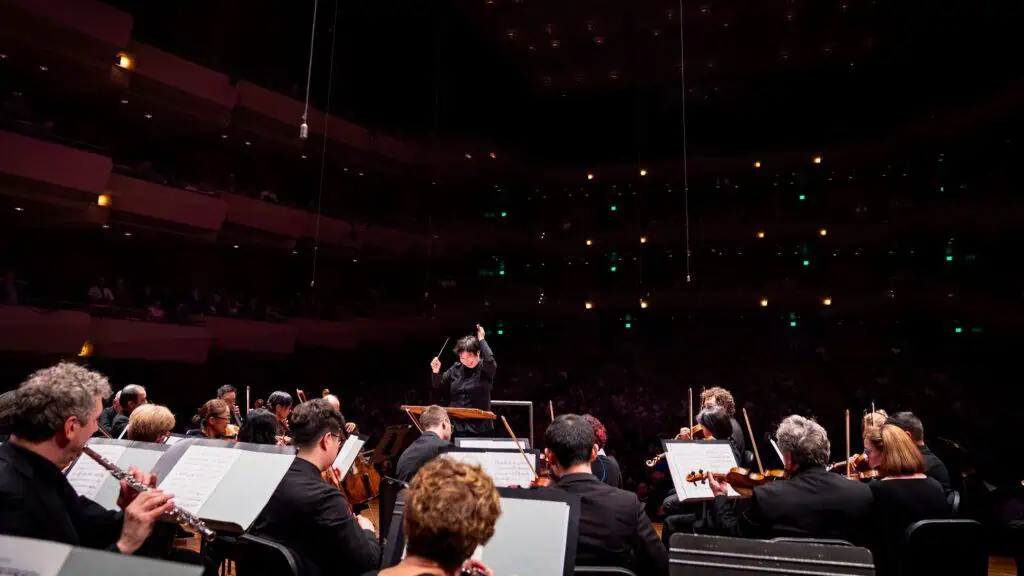A conductor leads an orchestra during a performance in a large concert hall, with musicians seated and audience members visible in the background.