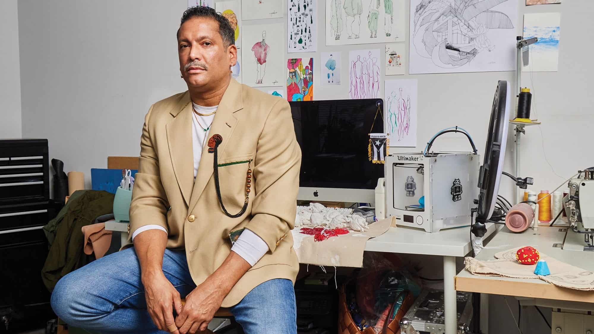 Seattle artist Gabriel-Bello Díaz sits on a desk in a studio filled with art supplies, tools, and sketches, evoking the creative energy found at King Street Station art spaces.