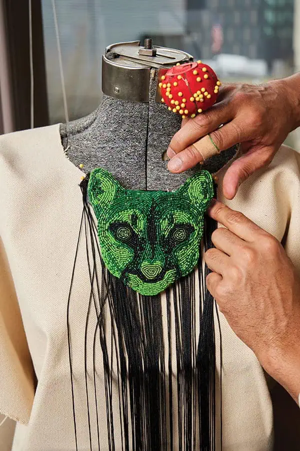 A Seattle artist adjusts a beaded green feline face with black fringe on a dress form at King Street Station art, wearing a red pincushion ring on their finger.