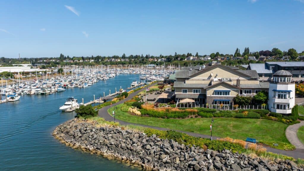 A sunny day over a harbor filled with sailboats and a large yacht. Buildings and green landscaping line the waterfront.