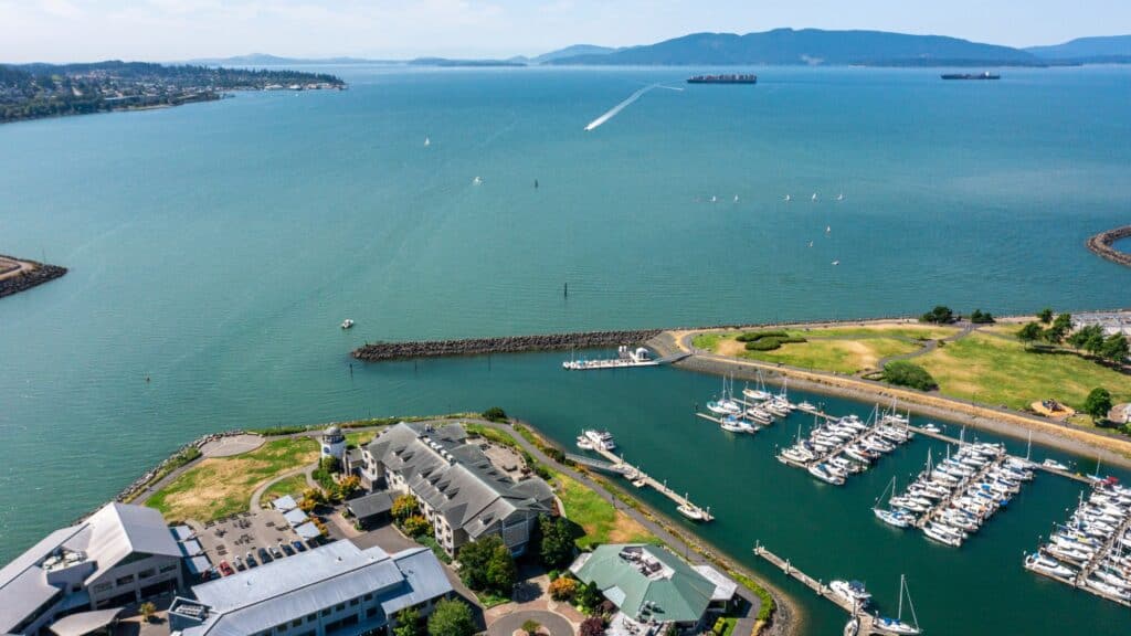 Aerial view of a marina with docked boats, a breakwater, and buildings near the shoreline, overlooking a large body of water with distant mountains.