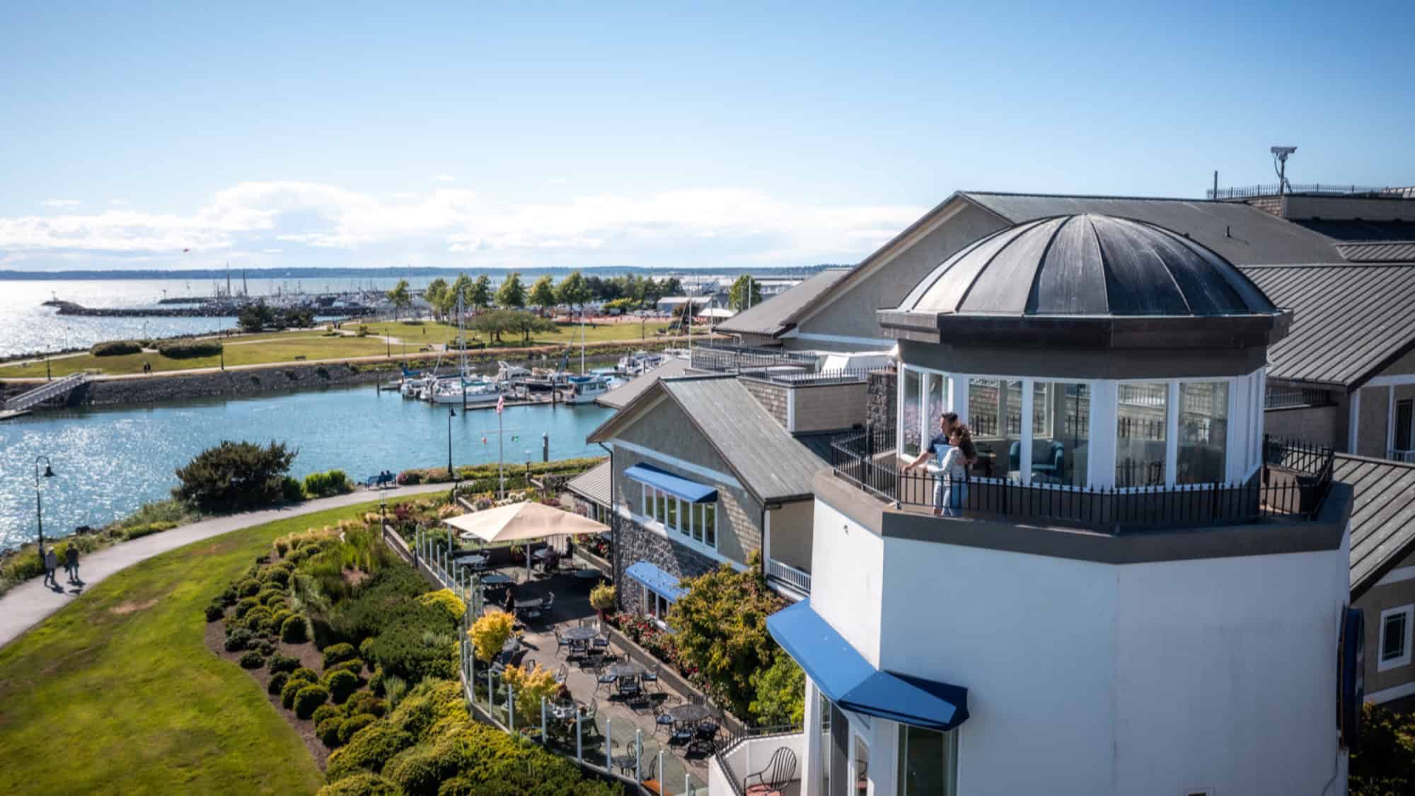 A waterfront building with a domed balcony overlooks a marina, landscaped gardens, outdoor seating, and a walking path beside the water under a clear blue sky.