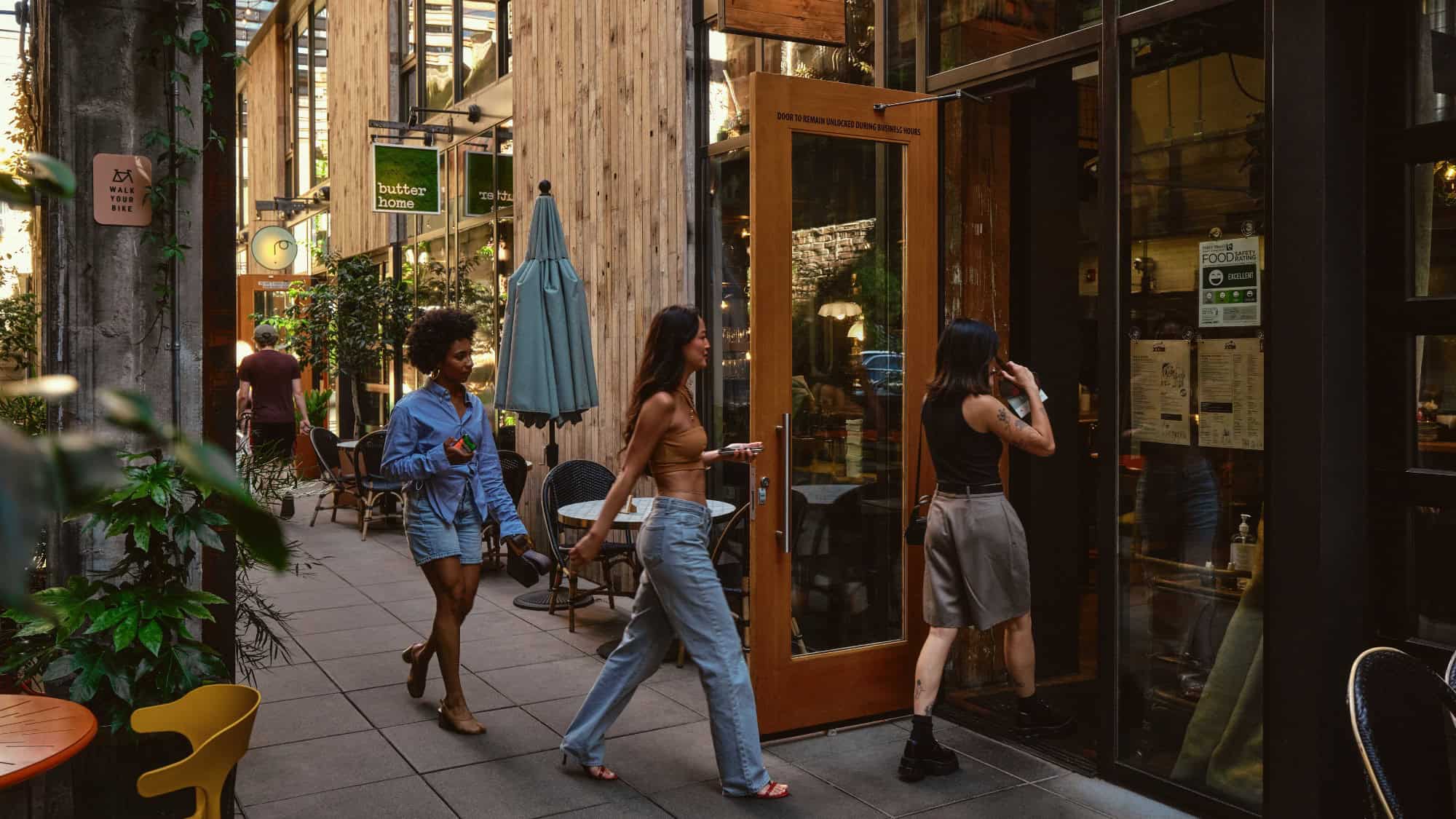 Three women are entering a café along a narrow outdoor walkway lined with tables, chairs, plants, and storefronts during the daytime.