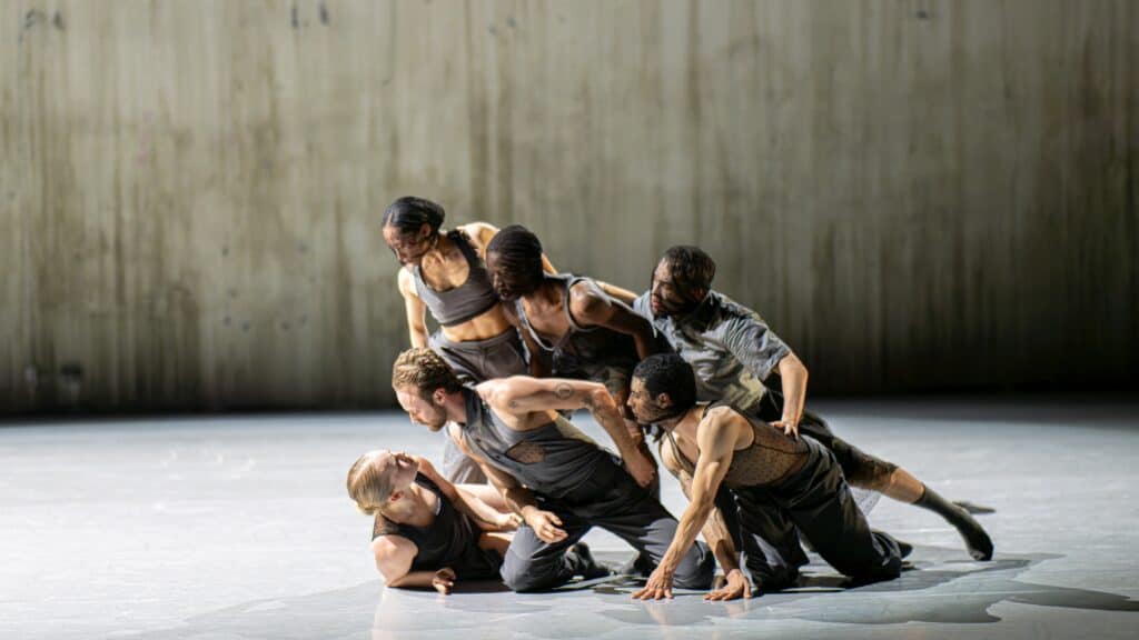 Six contemporary dancers in gray costumes pose closely together on a brightly lit stage with a plain, textured wall in the background.