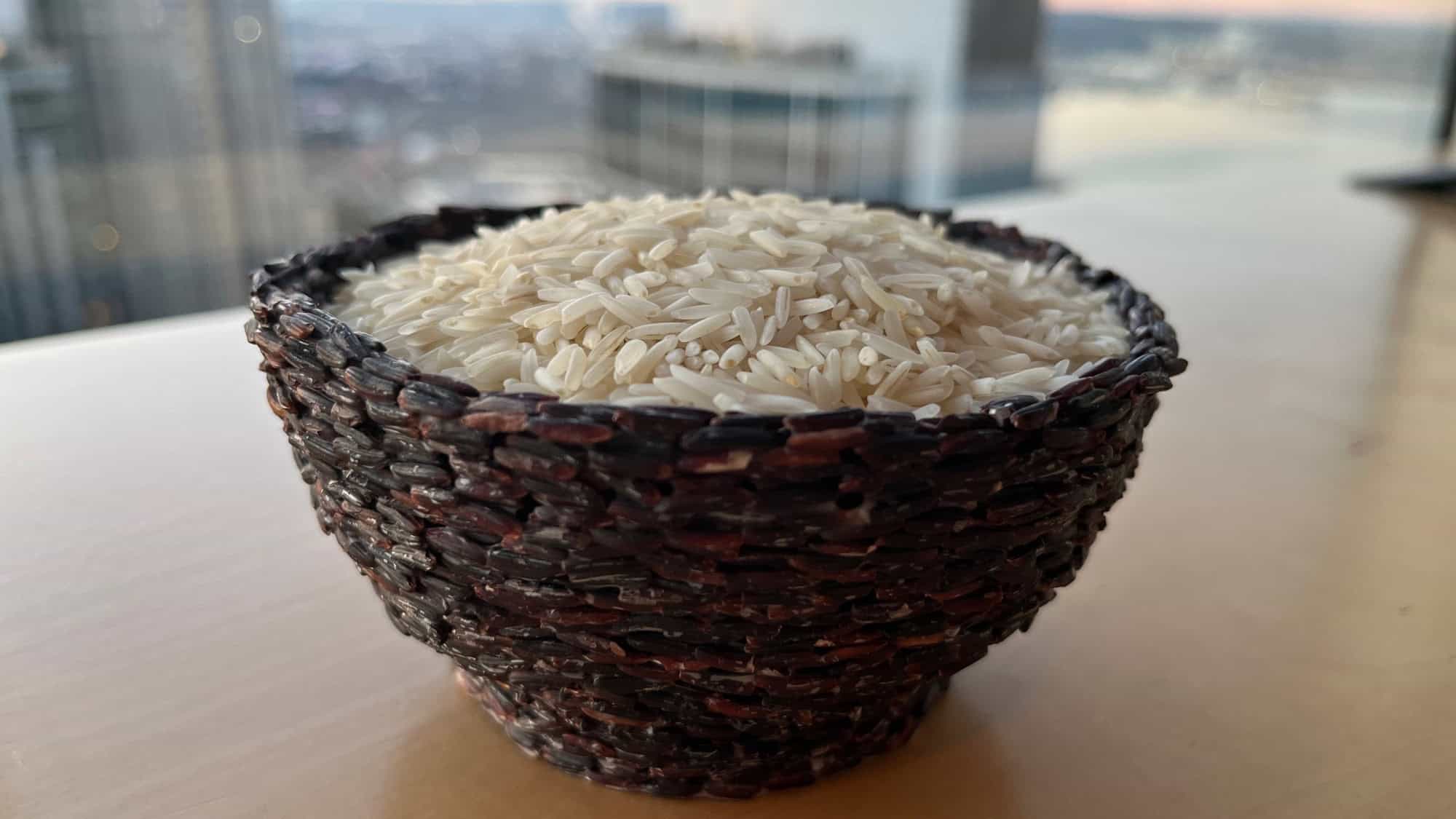 A woven dark brown bowl filled with uncooked white rice sits on a light-colored surface with city buildings in the blurred background.