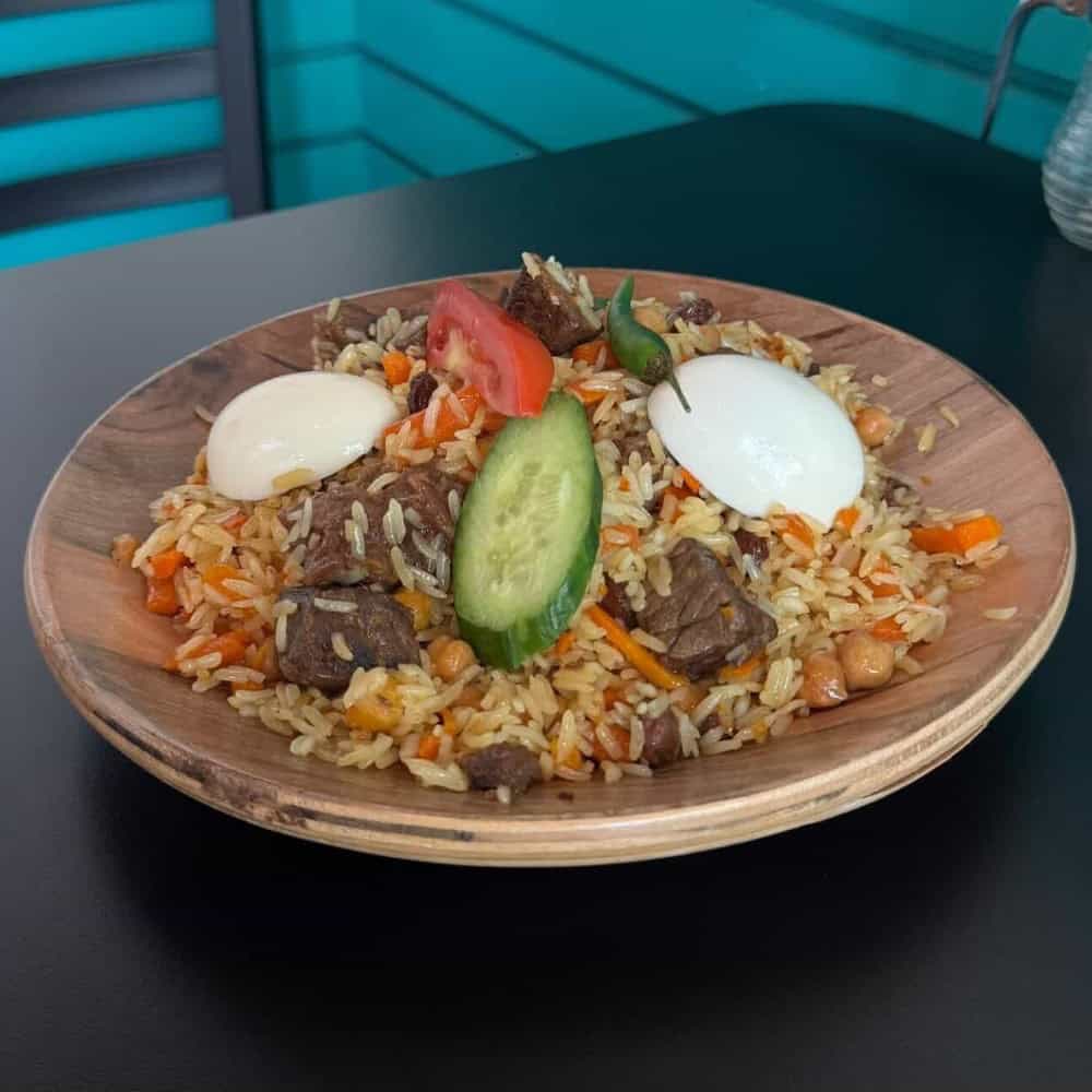 A wooden plate with rice, beef pieces, boiled eggs, carrots, chickpeas, a cucumber slice, tomato slice, and a green chili, on a dark table.