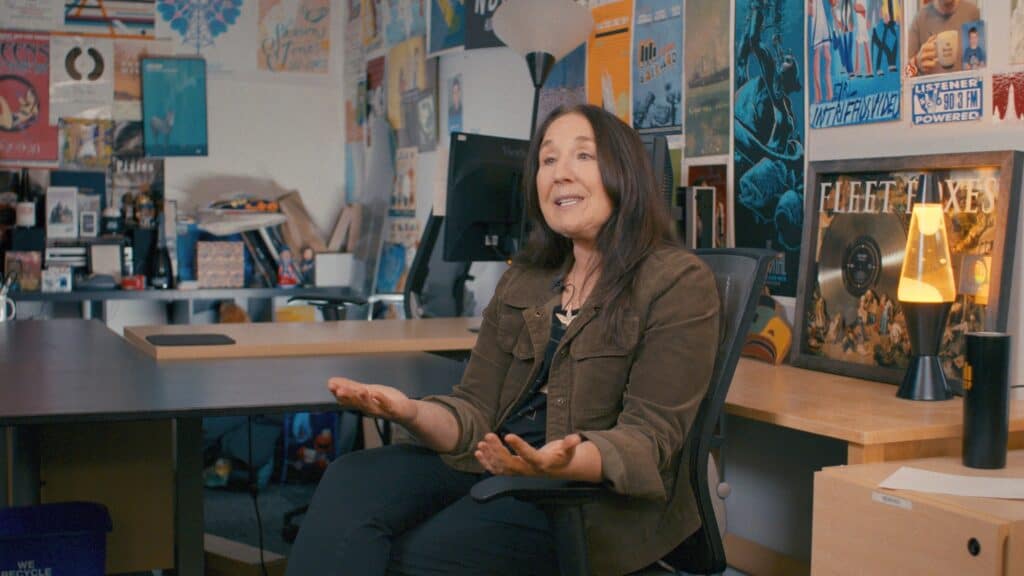 A woman sits in an office decorated with posters and albums, gesturing with open hands while speaking.