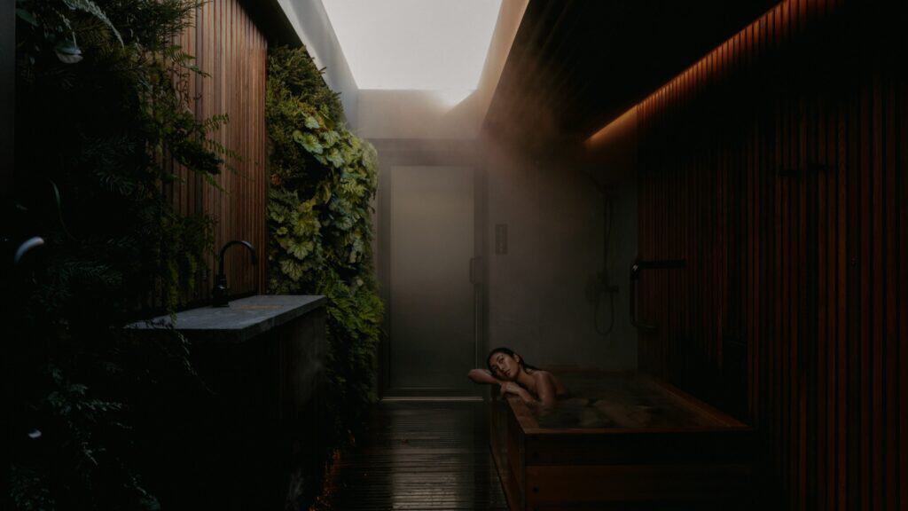 A person relaxes in a wooden soaking tub in a narrow, steamy room with vertical wooden slats and green plants on one wall.
