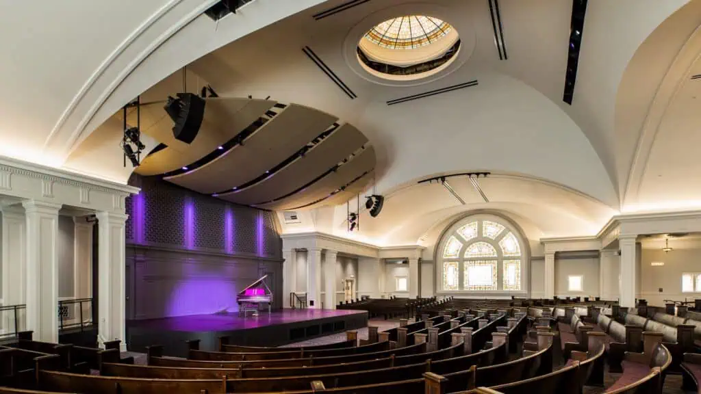 Wide view of an empty auditorium with wooden pews, a stage with a grand piano, arched windows, a dome skylight, and purple accent lighting.