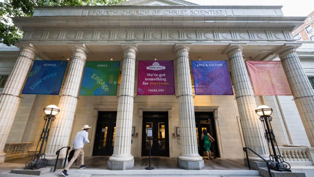 The entrance of a neoclassical building with four columns displays colorful banners labeled Science, Civics, Arts & Culture, and Community. People are entering the building.