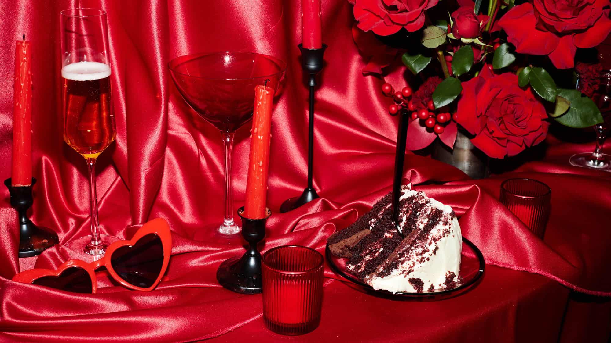 A slice of chocolate cake, red candles, champagne, heart-shaped sunglasses, and a bouquet of red roses arranged on red satin fabric.
