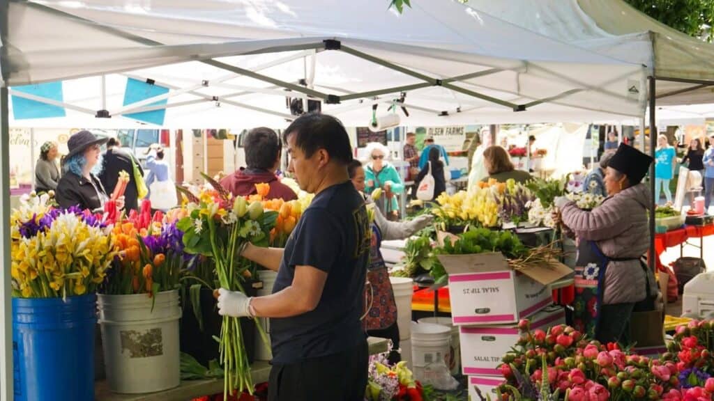 A person arranges flowers at an outdoor market stall under a white canopy, surrounded by colorful flower bouquets and other shoppers enjoying spring in Seattle.