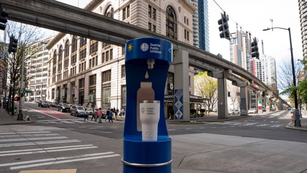 A Seattle Public Utilities water refill station stands on a city sidewalk near a street intersection with crosswalks and surrounding buildings.