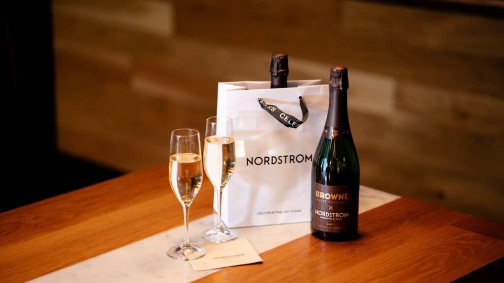 A bottle of Browne x Nordstrom sparkling wine, two filled champagne flutes, and a Nordstrom gift bag on a wooden table.