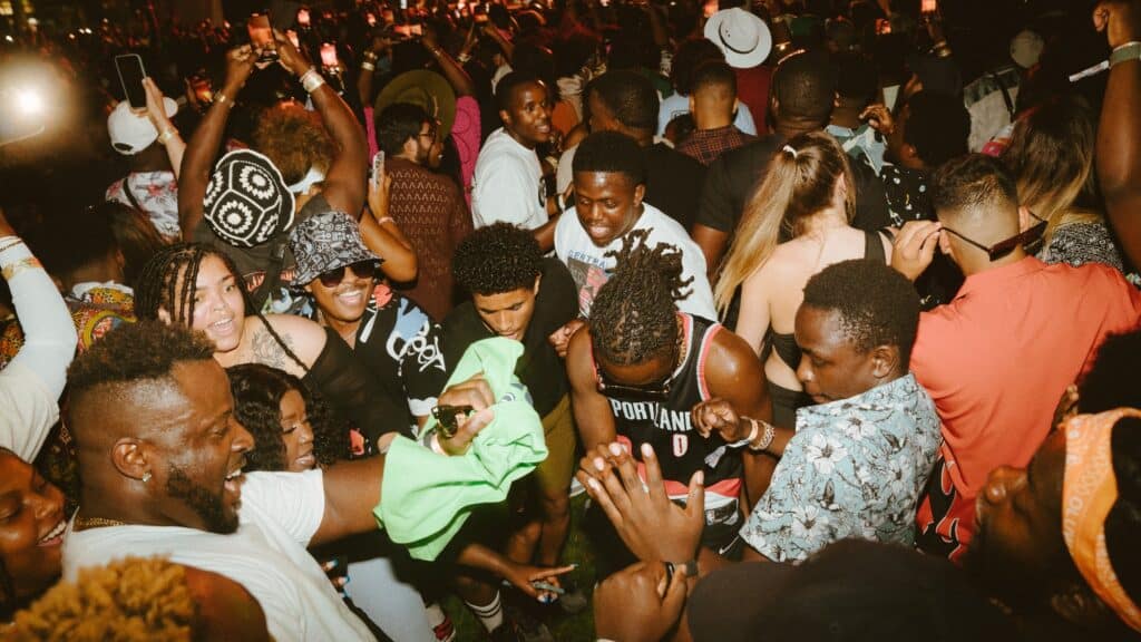 A large group of people gather closely together outdoors at night on the Seattle waterfront, dancing and smiling in a lively BLASTFEST party atmosphere, celebrating Spring in the Pacific Northwest.