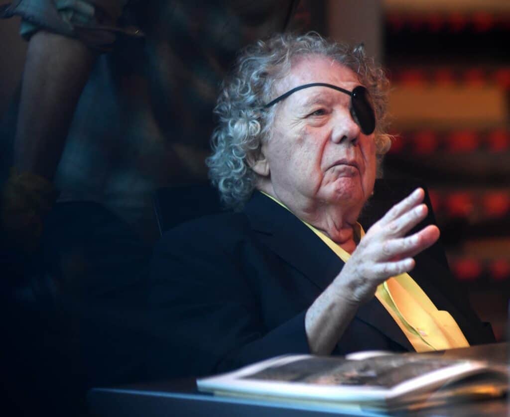 An older man with curly hair and an eye patch sits at a table with an open book, gesturing with his hand.