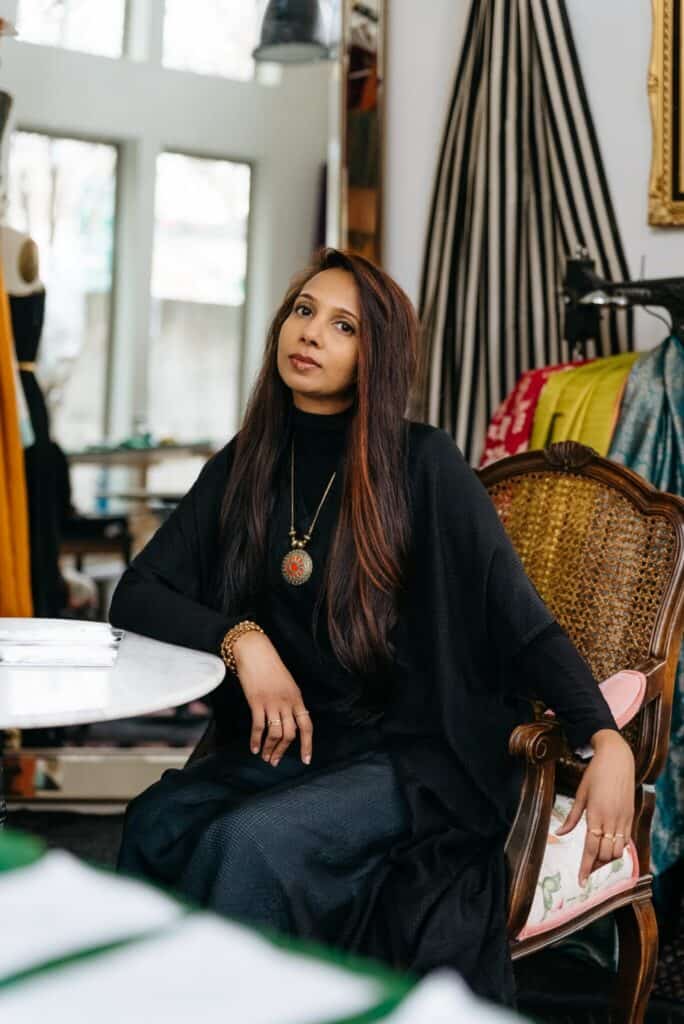 A woman with long hair, dressed in black and wearing a pendant necklace, sits on a cane chair in a stylish, well-lit room with fabric and clothing in the background.