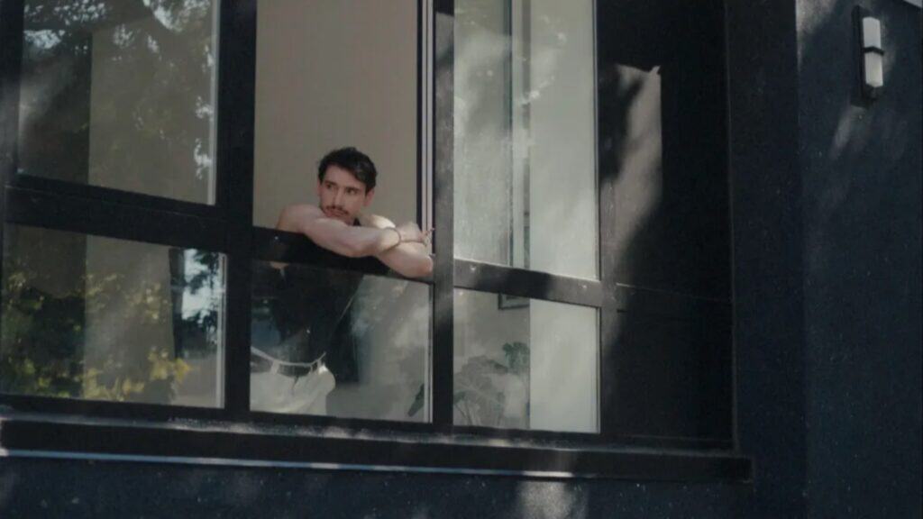 A man leans on a windowsill, looking outside through an open window of a dark-colored building, reflecting on the SIFF movies lineup.