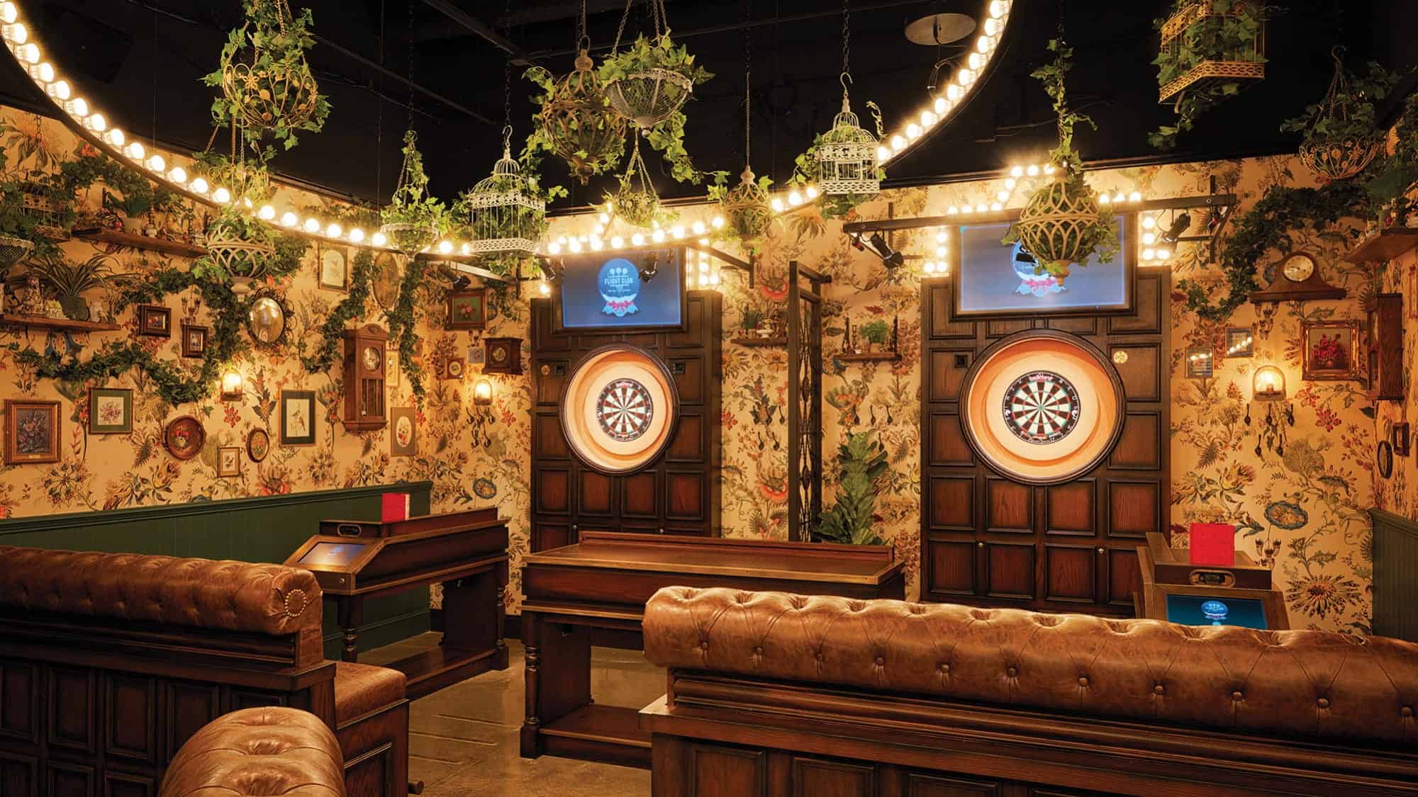 A stylish dart room with brown leather couches, floral wallpaper, two dartboards, hanging plants, decorative lights, and a curated wine selection from Offset Bottle Shop for those who value ethical practices.