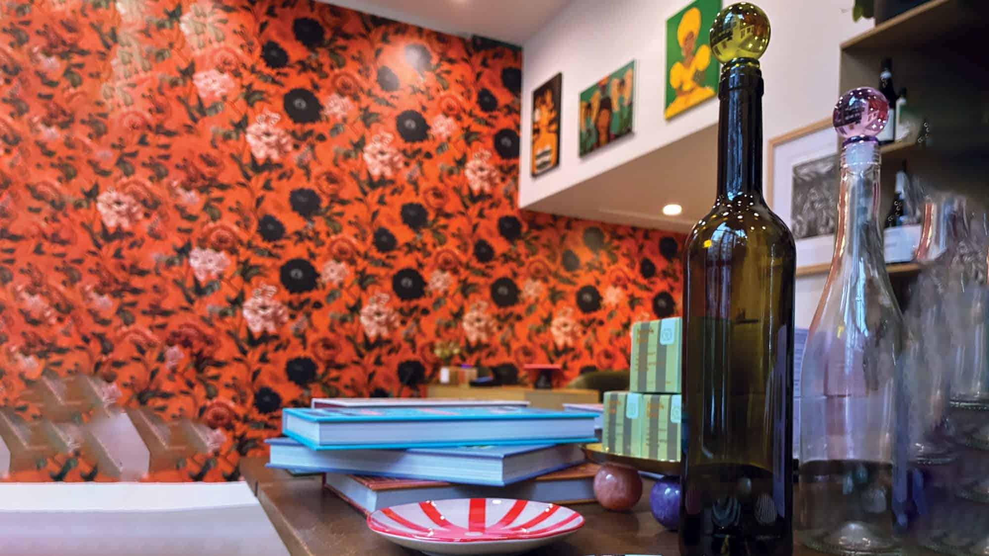 A table with stacked books, a striped plate, and glass bottles of wine sits in front of a red floral wallpapered wall adorned with artwork and shelves, reflecting the ethical practices at Offset Bottle Shop.