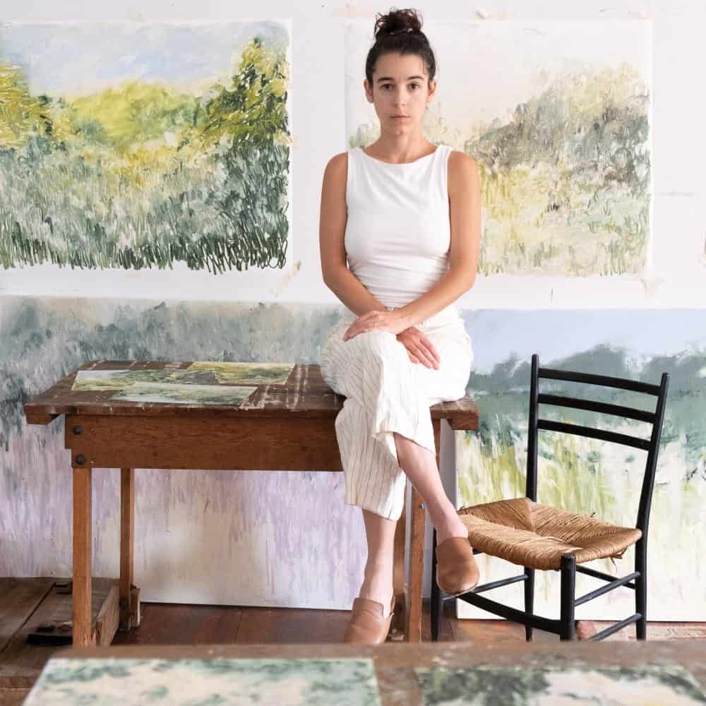 A woman in a white top and light pants sits on a wooden table in the gallery space at Dirkse/Prim Gallery, with landscape paintings behind her and a black chair beside her.