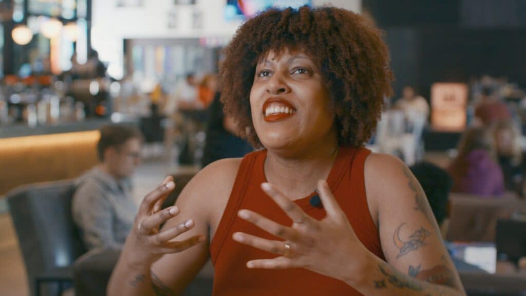 A person with curly hair and tattoos, wearing a sleeveless red top, gestures expressively while speaking in a busy indoor setting.