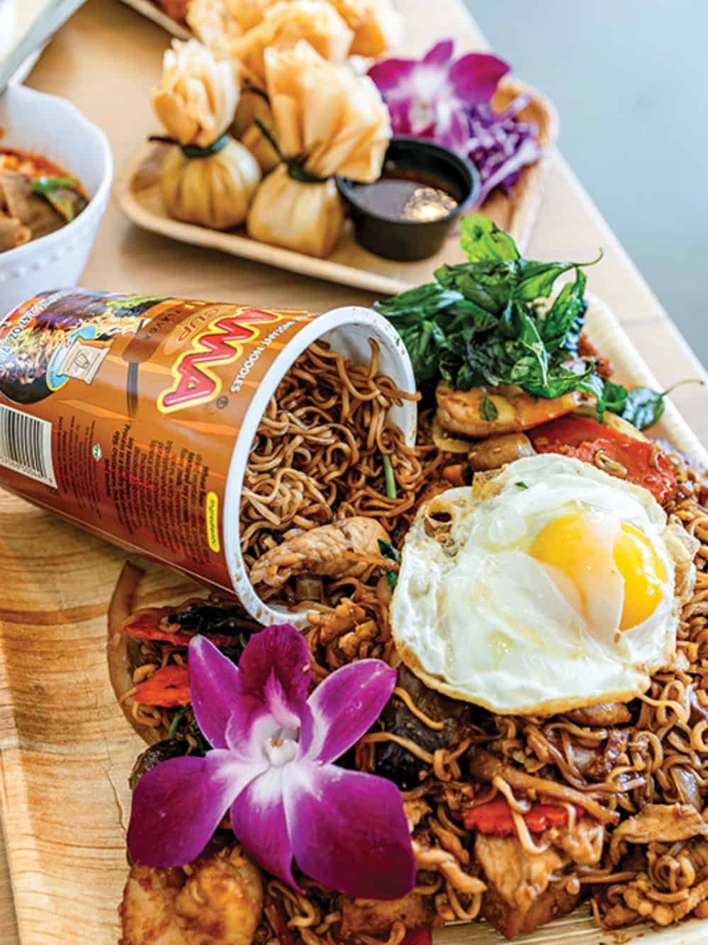 A plate of stir-fried noodles with vegetables, chicken, and a fried egg is served at Bloom Thai in Lakewood, adorned with an orchid flower and instant noodle cup, plus crispy fried dumplings with dipping sauce in the background.