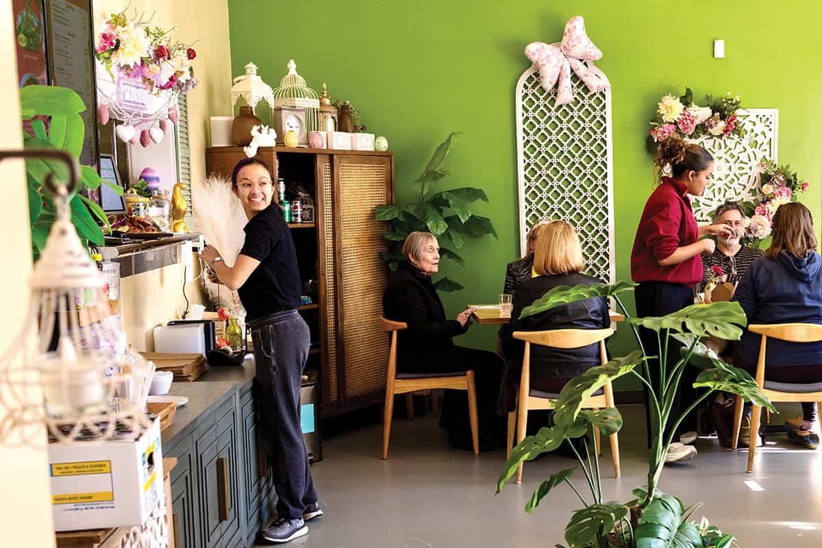 A woman smiles while cleaning near a group of people chatting at Bloom Thai, a Seattle fusion Thai cafe in Lakewood, beautifully decorated with plants and floral arrangements.