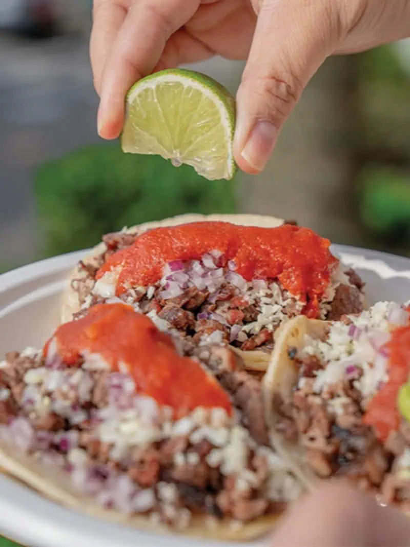 A person squeezes a lime wedge over three street-style beef tacos topped with diced onion, cilantro, and red salsa on a white plate—an inviting scene for anyone exploring new restaurants in Seattle.