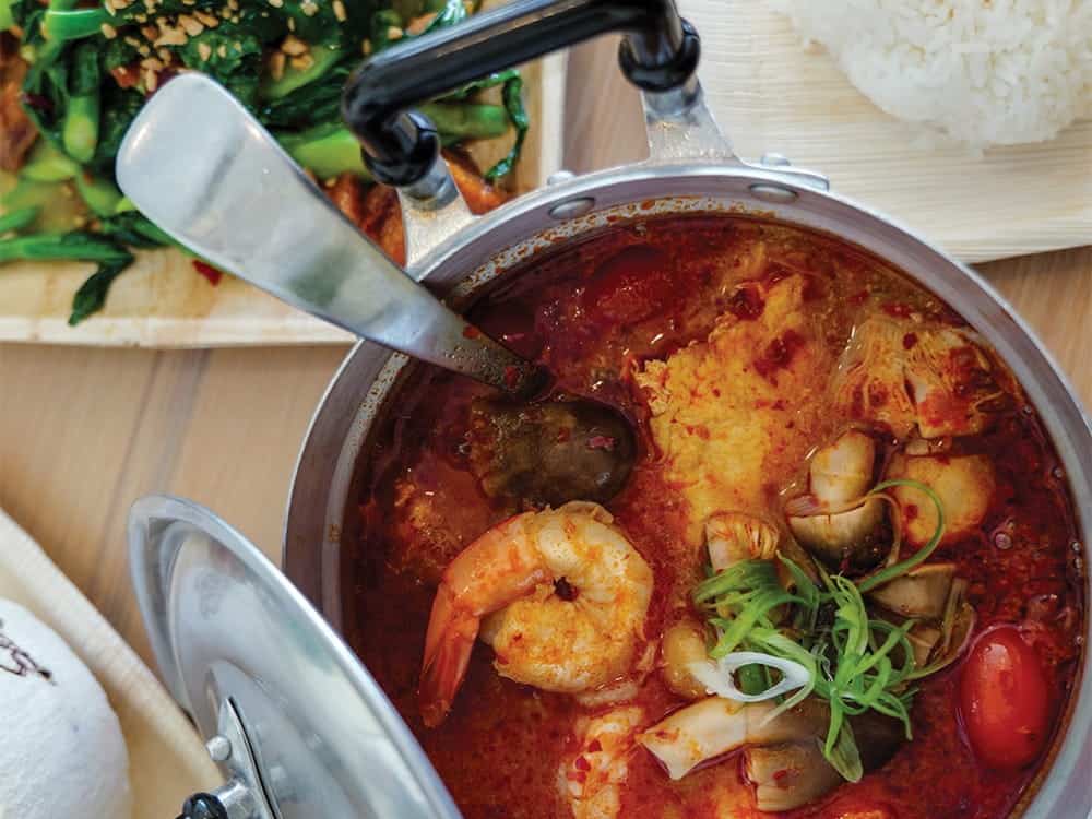 A pot of spicy seafood soup with shrimp, mushrooms, tomatoes, and green onions, served alongside rice and sautéed greens—a delicious taste of Seattle fusion Thai from Bloom Thai.