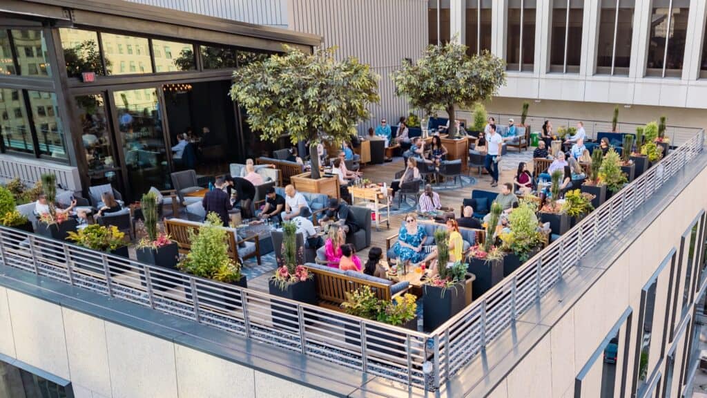 A rooftop restaurant with several groups of people sitting at tables, surrounded by plants and trees, enjoying food and drinks on a sunny day.