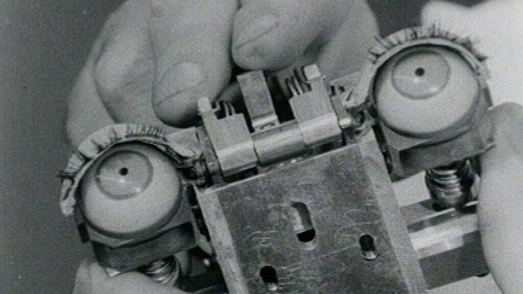 Close-up of hands holding a mechanical device resembling a pair of eyes, with eyelashes, metal springs, and gears visible.