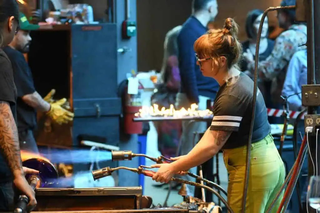 A woman uses a blowtorch to shape glass in a workshop, with other people working and observing in the background.
