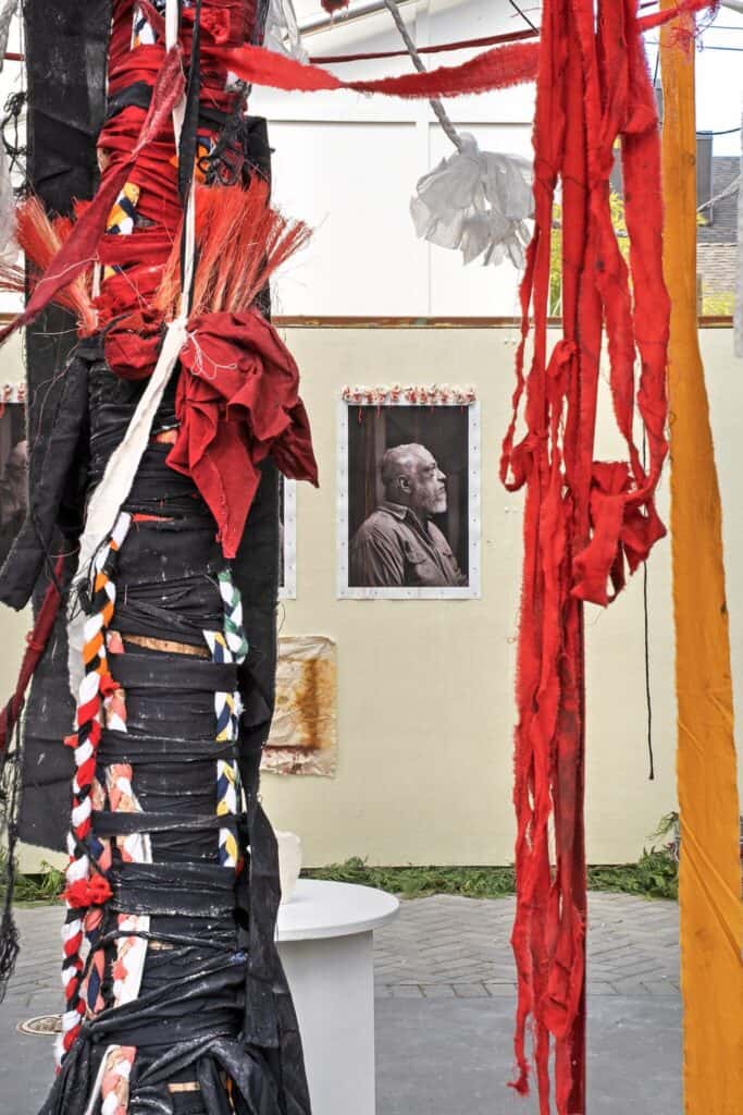 Large poles wrapped in black and red fabric strips stand in front of a wall displaying black-and-white portraits of an elderly man.