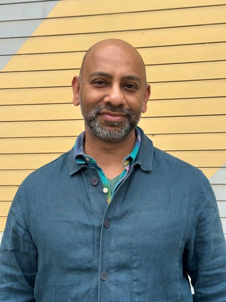 A man with a beard and shaved head wearing a teal jacket and green shirt stands in front of a yellow and gray striped wall.