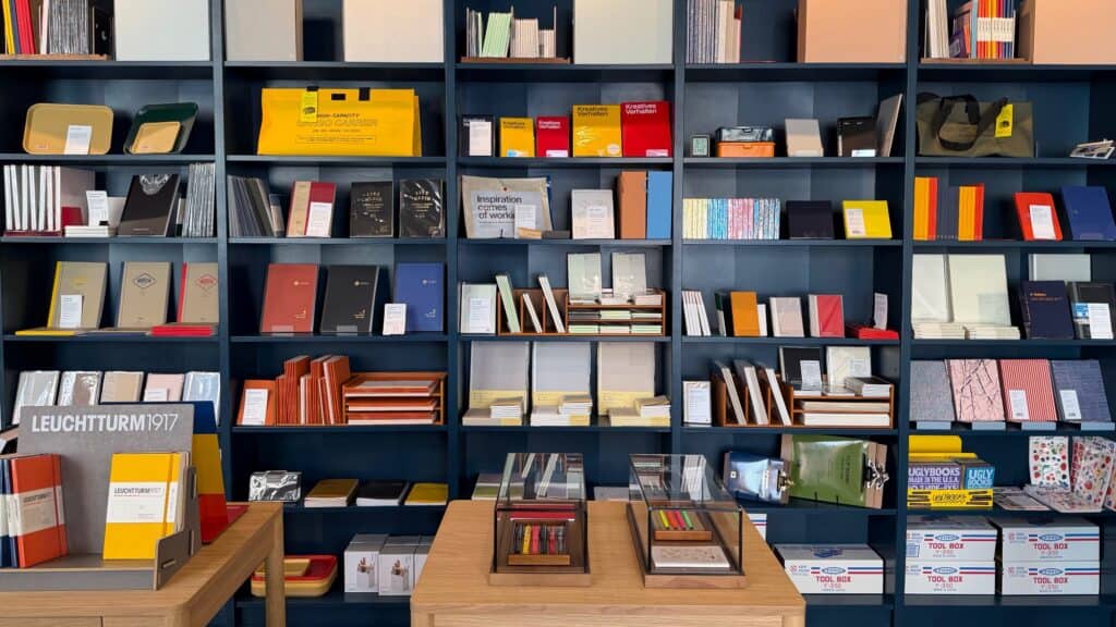 Shelves filled with various notebooks, stationary items, and office supplies are displayed in a store with wooden tables in the foreground.