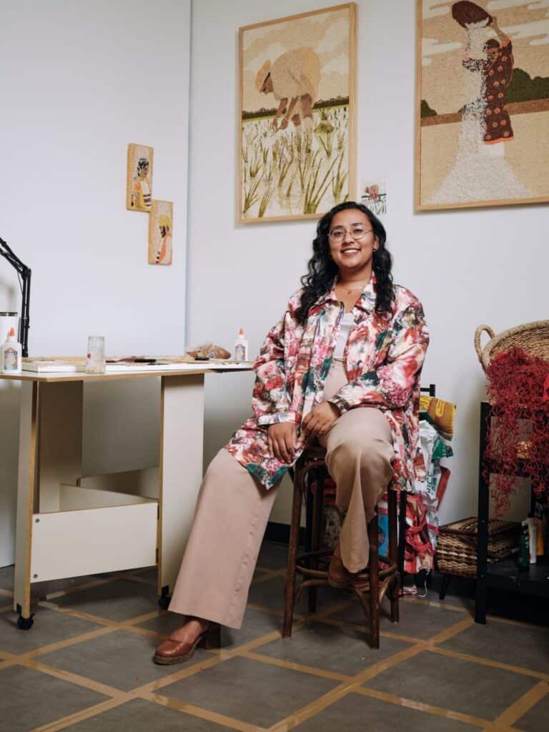 A woman wearing a floral coat sits on a stool in an art studio, surrounded by artwork, crafting materials, and a woven basket.