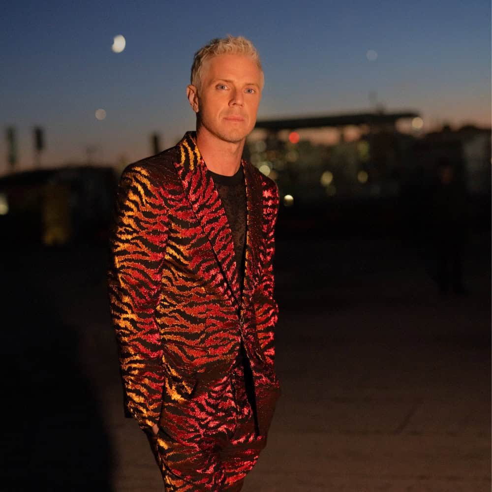A man with short blond hair wears a red and yellow patterned suit, standing outdoors at dusk with a cityscape and crescent moon in the background.