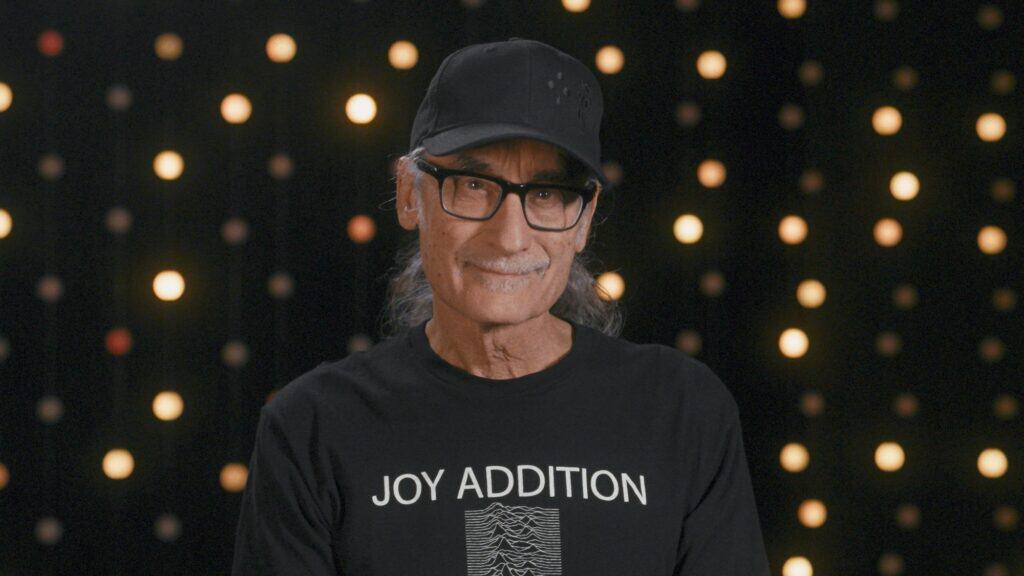 An older man with long gray hair, glasses, and a black cap stands in front of a backdrop with small lights, wearing a black "Joy Addition" t-shirt.