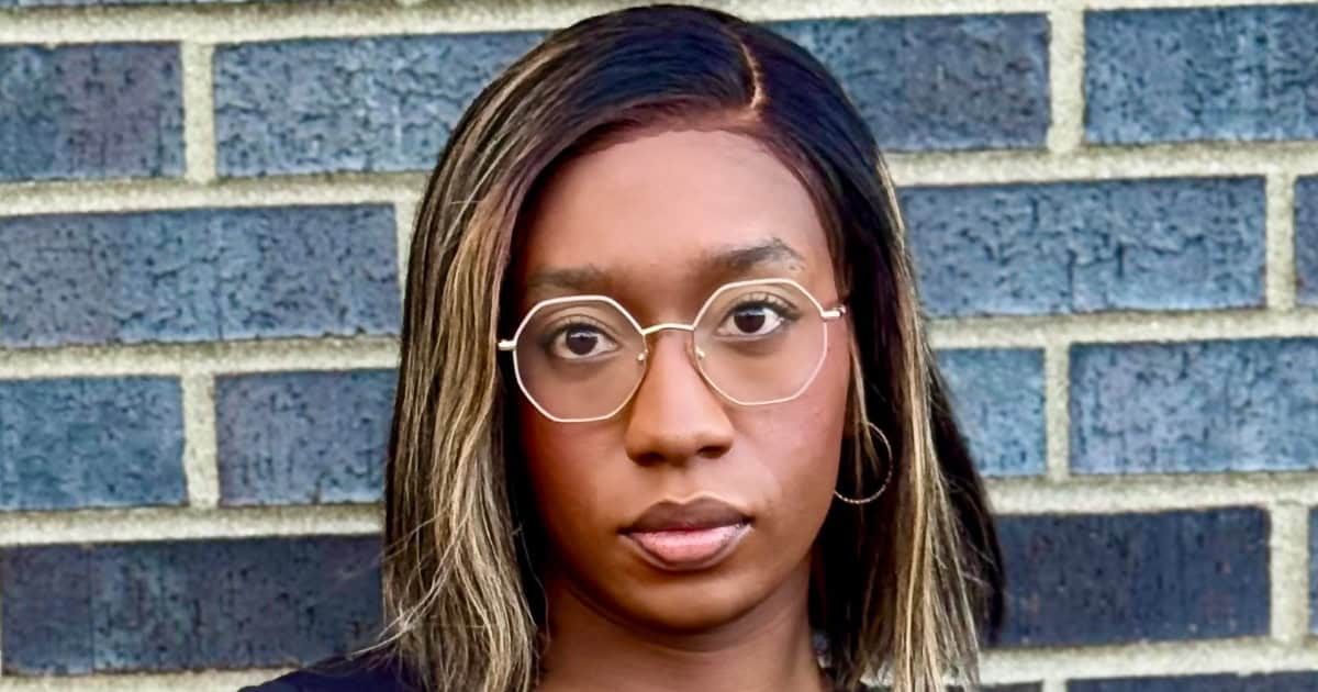 A person with shoulder-length hair and round glasses stands in front of a brick wall, looking directly at the camera.